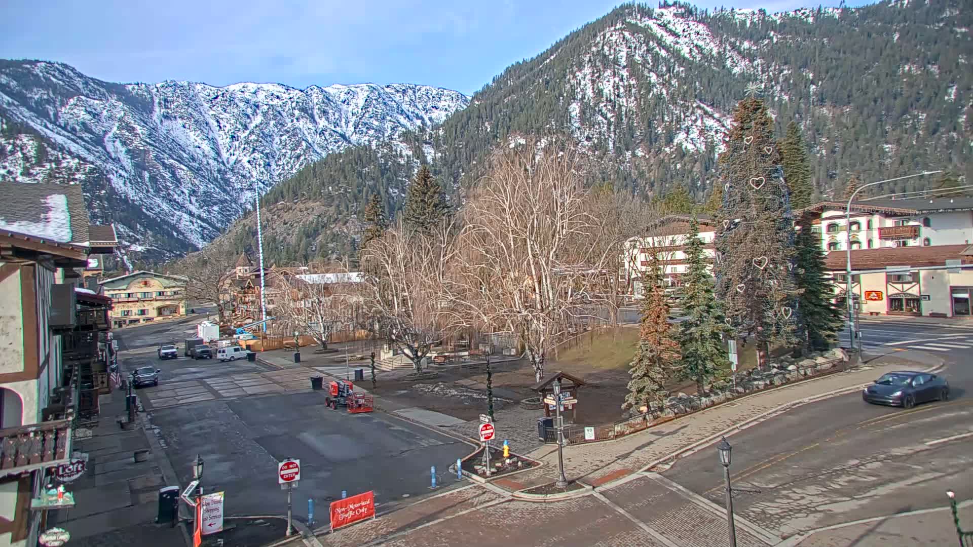 A sunny day in a mountain town shows a sparsely populated town square with a decorated evergreen tree, a few vehicles, and snow-capped mountains in the background.