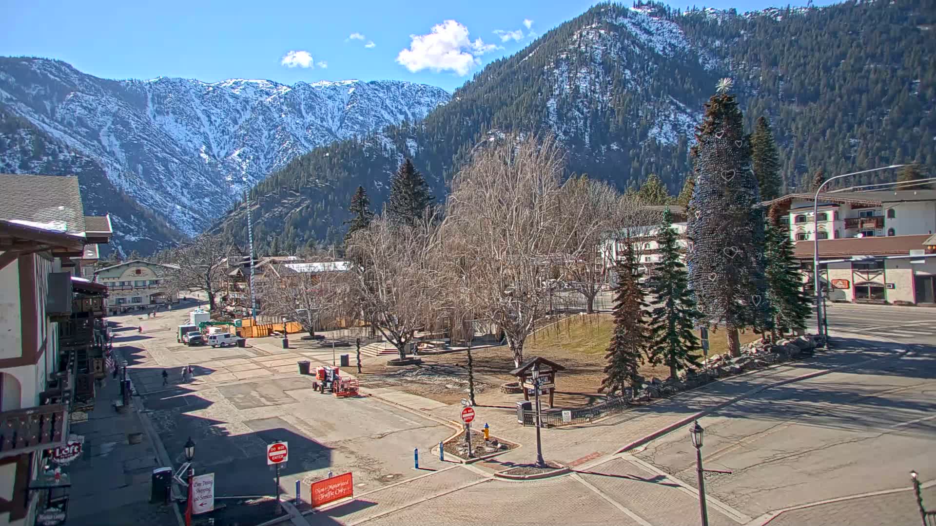 A mostly empty town square sits in front of snow-capped mountains on a sunny day, with a large artificial Christmas tree in the center.