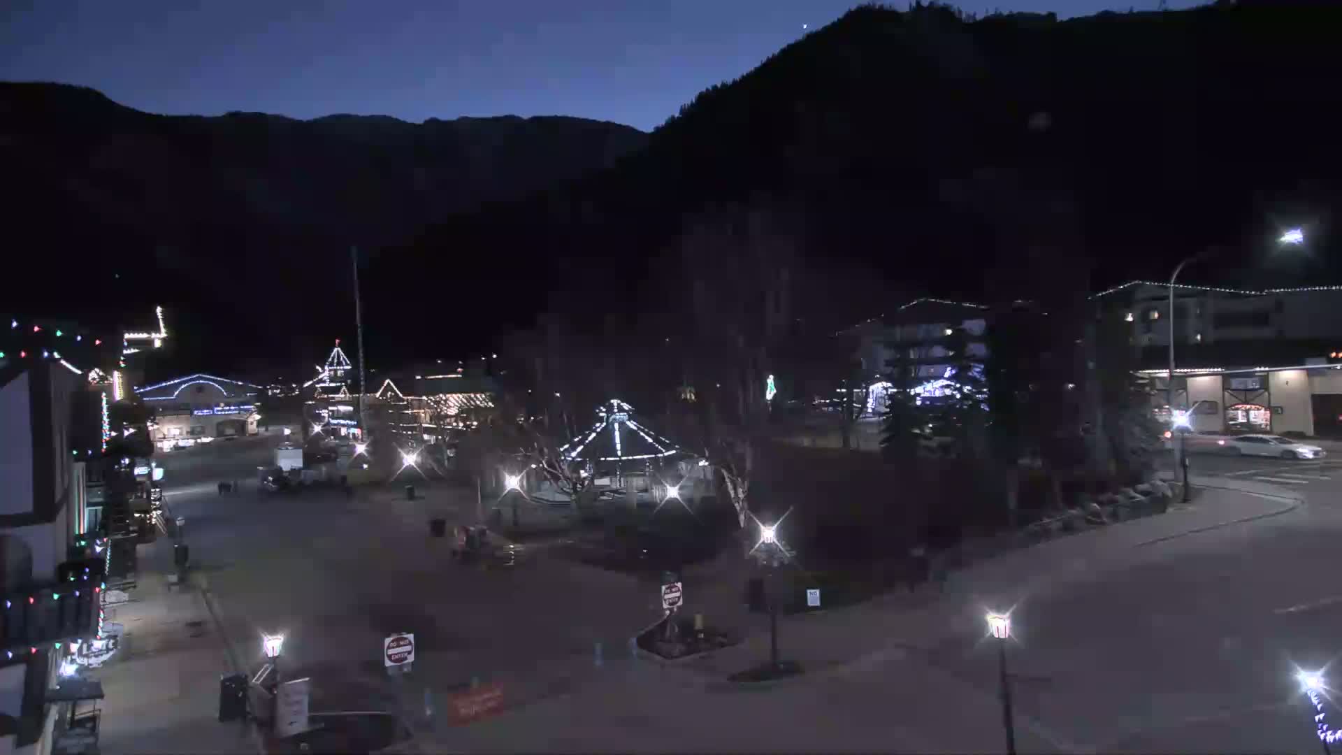 A nighttime view of a town square illuminated with Christmas lights, nestled against a dark mountain under a clear, dark sky.
