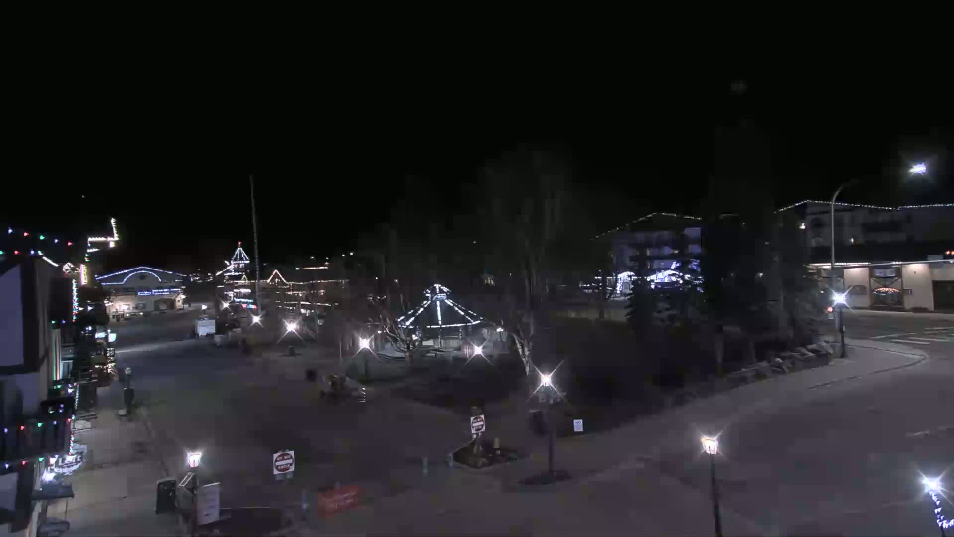 A nighttime view of a town square with buildings decorated with lights, a gazebo, and streetlights illuminating the mostly empty streets under a clear, dark sky.