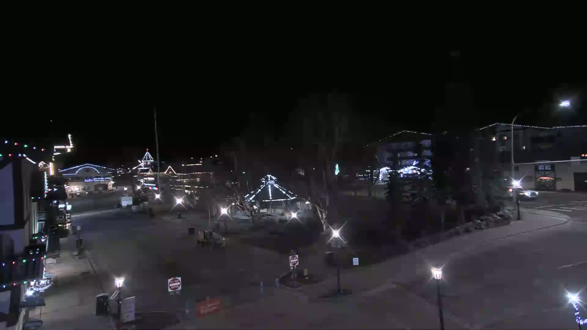 A nighttime view of a town square with festively lit buildings, a carousel, and lampposts, under a clear, dark sky.