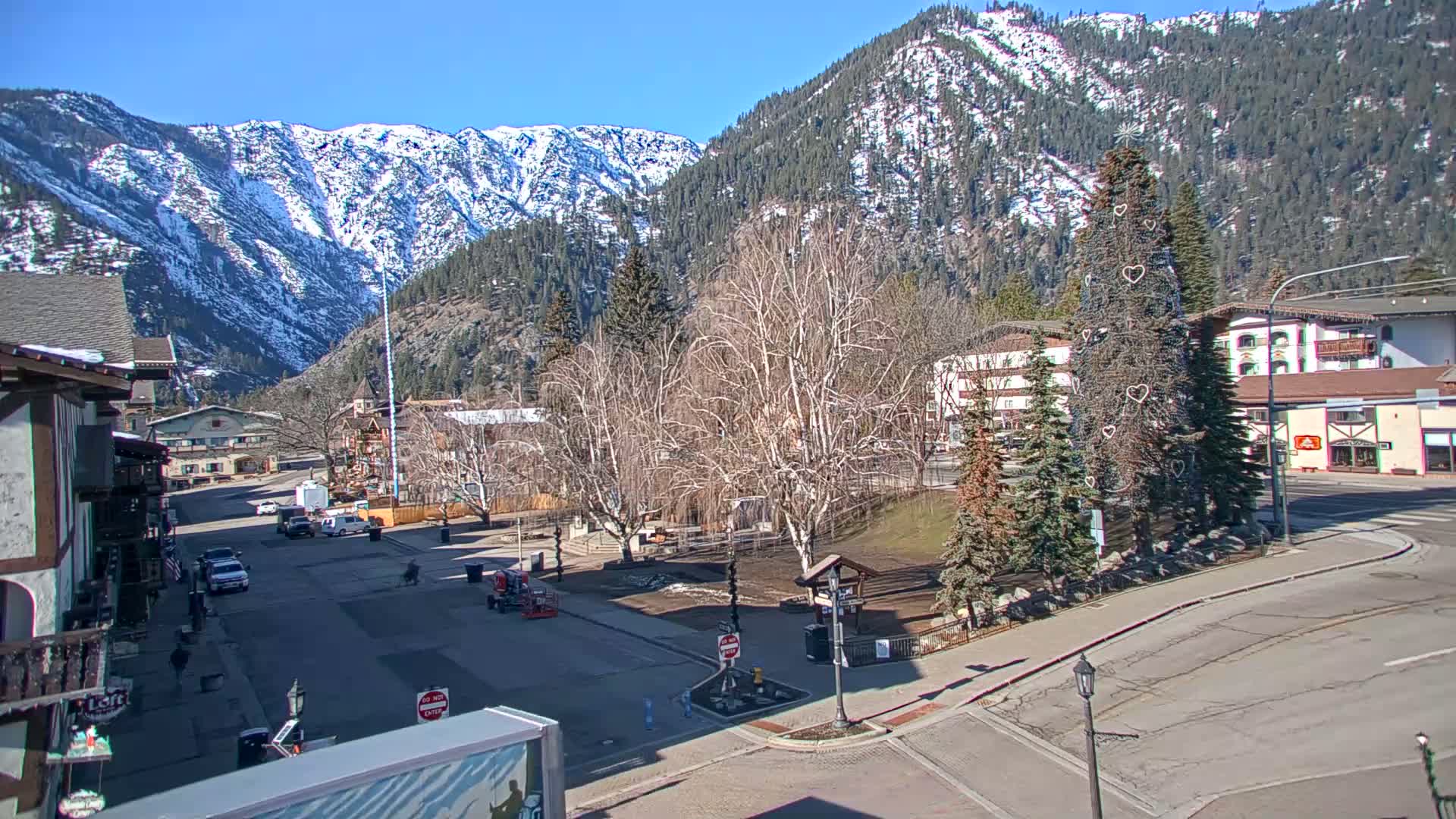 A mostly empty town square with bare trees and a decorated Christmas tree is situated in front of buildings, with snow-capped mountains in the background under a sunny sky.
