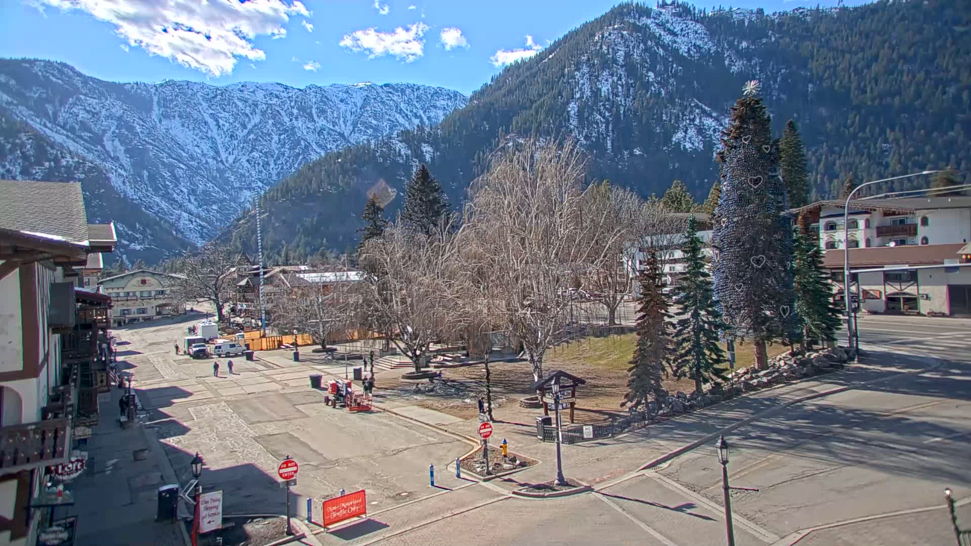 A sunny day reveals a mountain town square with a decorated evergreen tree, bare deciduous trees, and snow-capped mountains in the background.