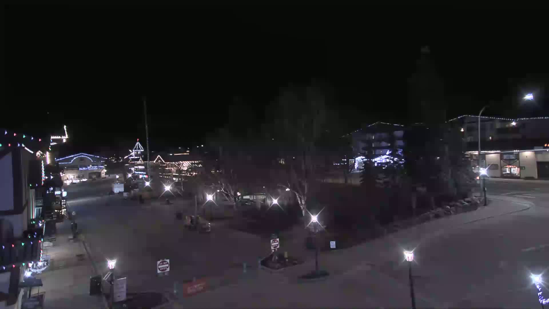 A nighttime view of a town square with buildings decorated with Christmas lights, under a clear, dark sky.