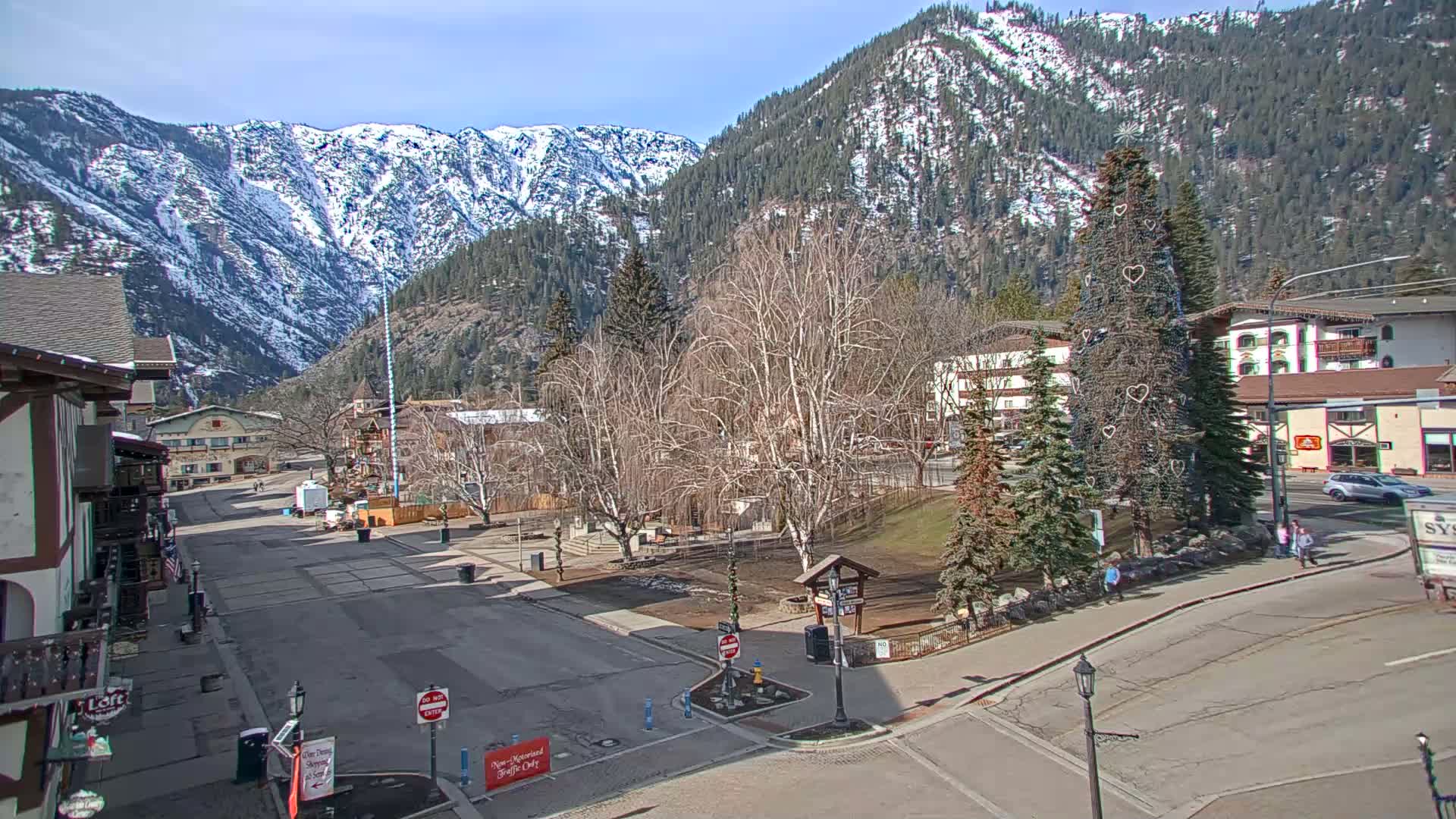 A mostly empty town square, surrounded by buildings and snow-capped mountains under a sunny sky, features a decorated evergreen tree in the center.