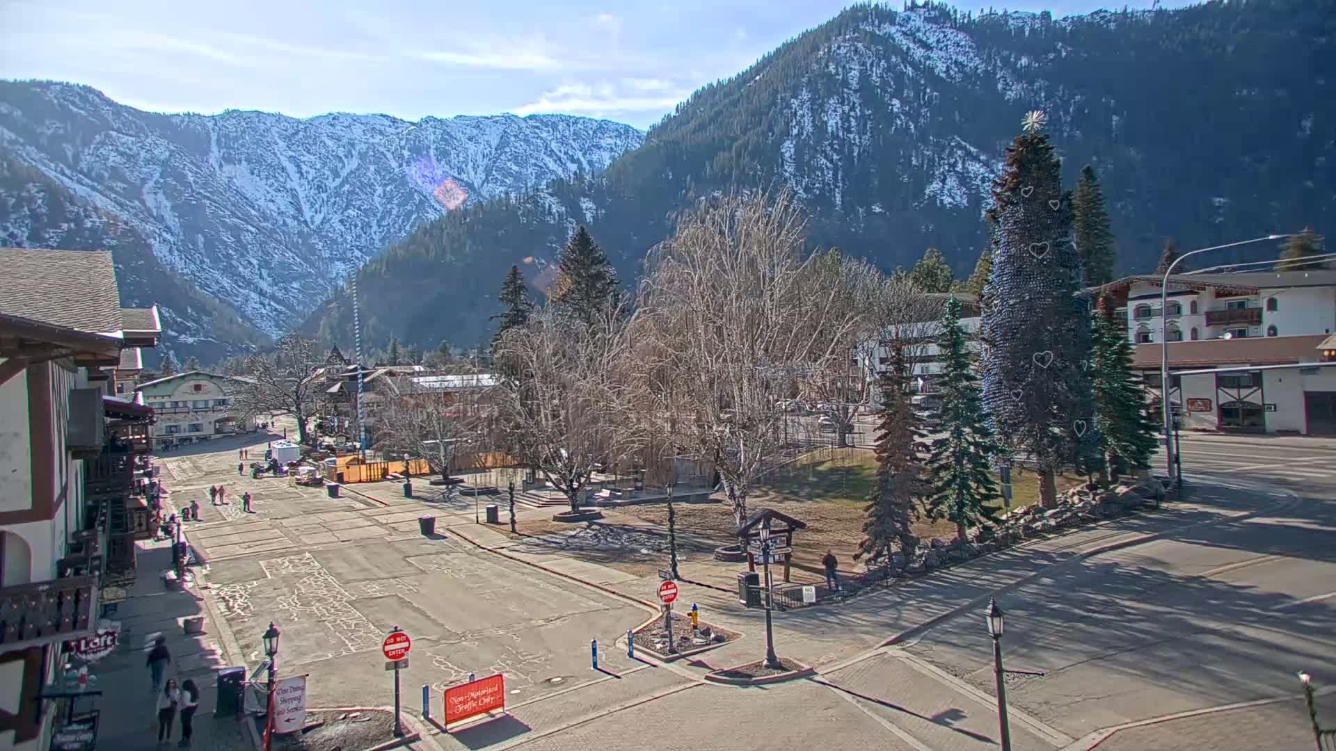 A sunny day in a snow-capped mountain town shows a paved town square with a large decorated evergreen tree in the center, surrounded by buildings and bare deciduous trees.