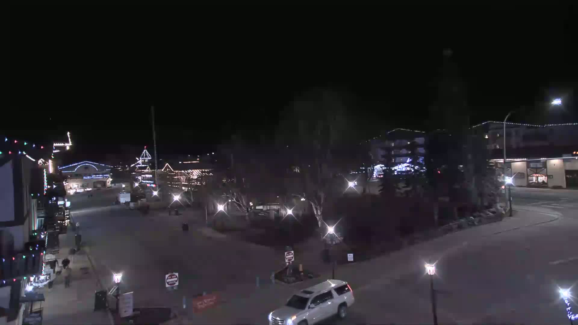 A nighttime view of a town square with several buildings decorated with lights, a few cars, and sparsely populated streets under a clear, dark sky.