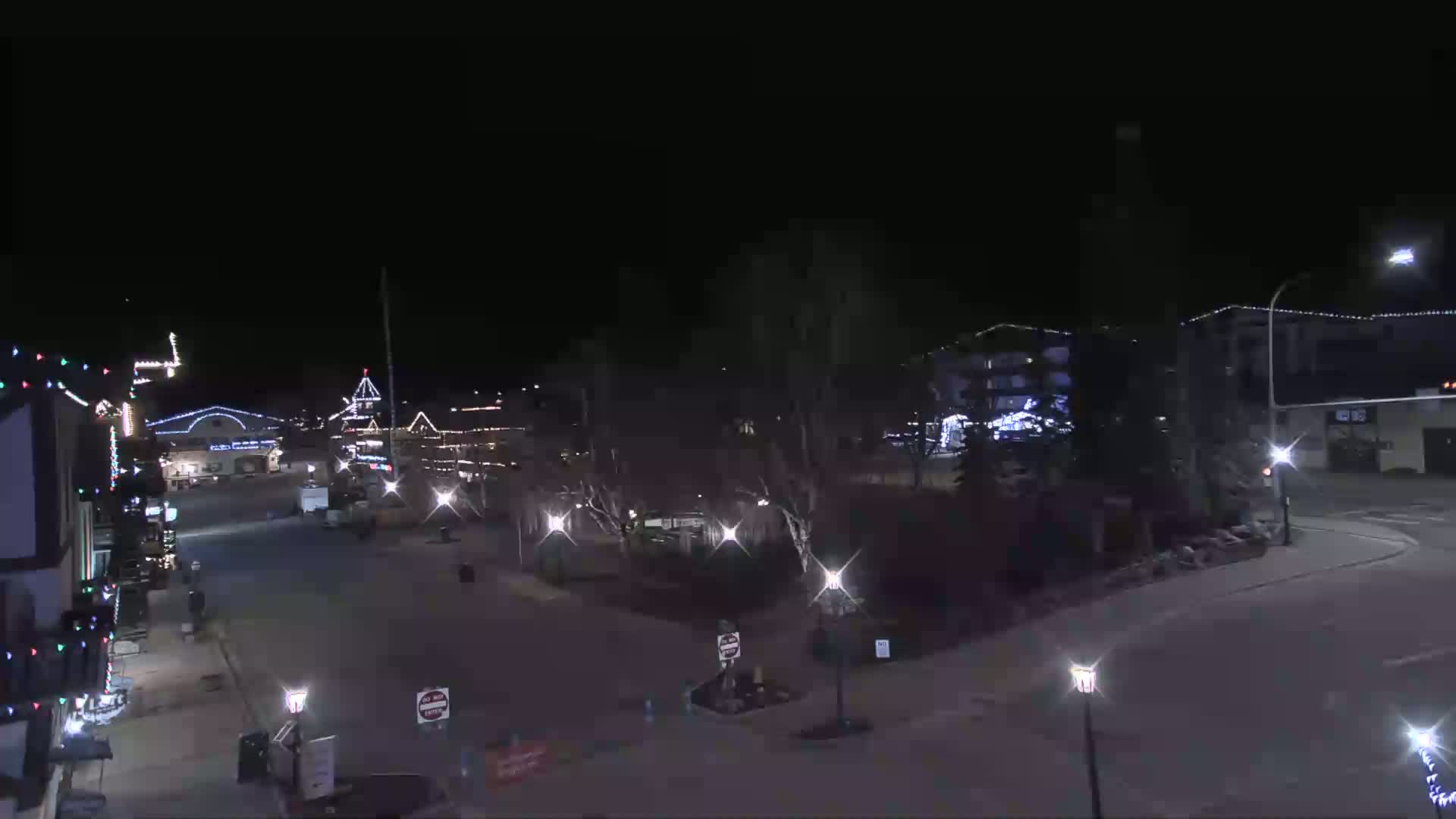 A nighttime view of a town square with several buildings decorated with lights, trees, and street lamps illuminating the mostly empty streets under a clear, dark sky.