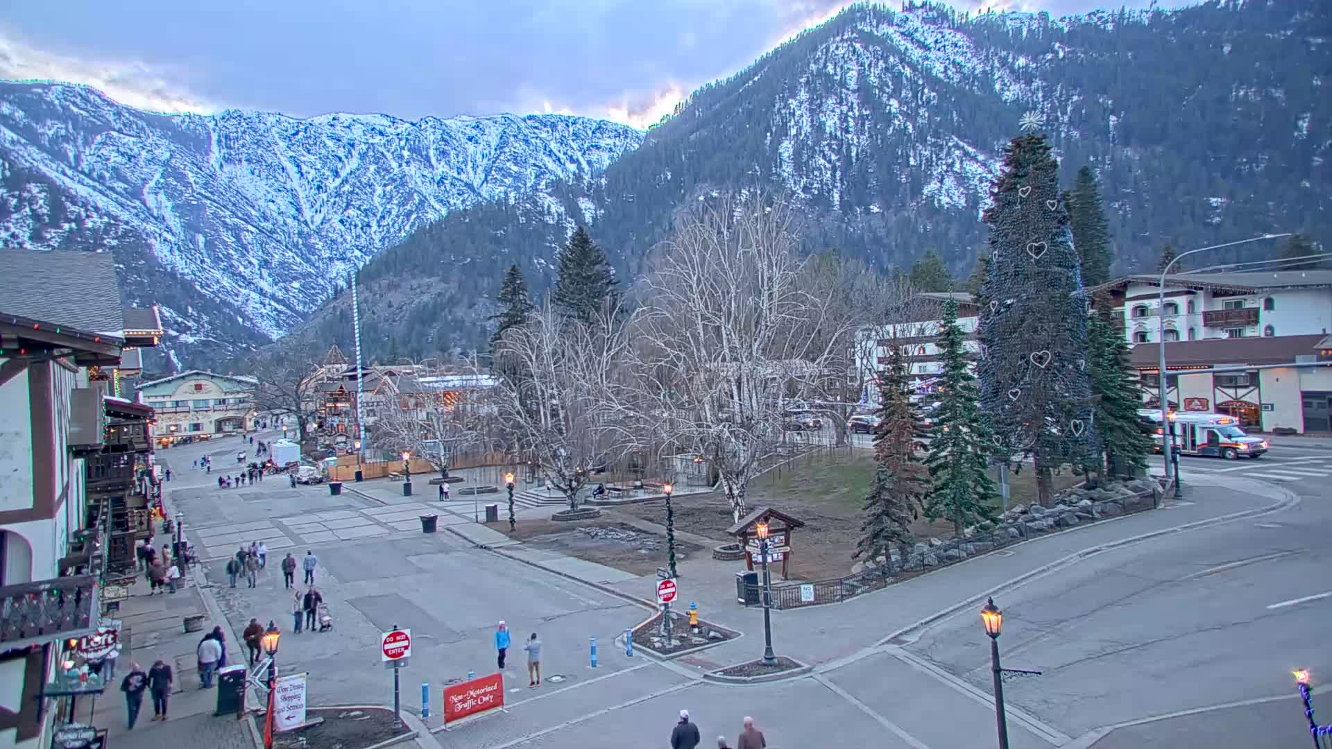 A town square with pedestrians, surrounded by buildings and snow-covered mountains under a twilight sky.