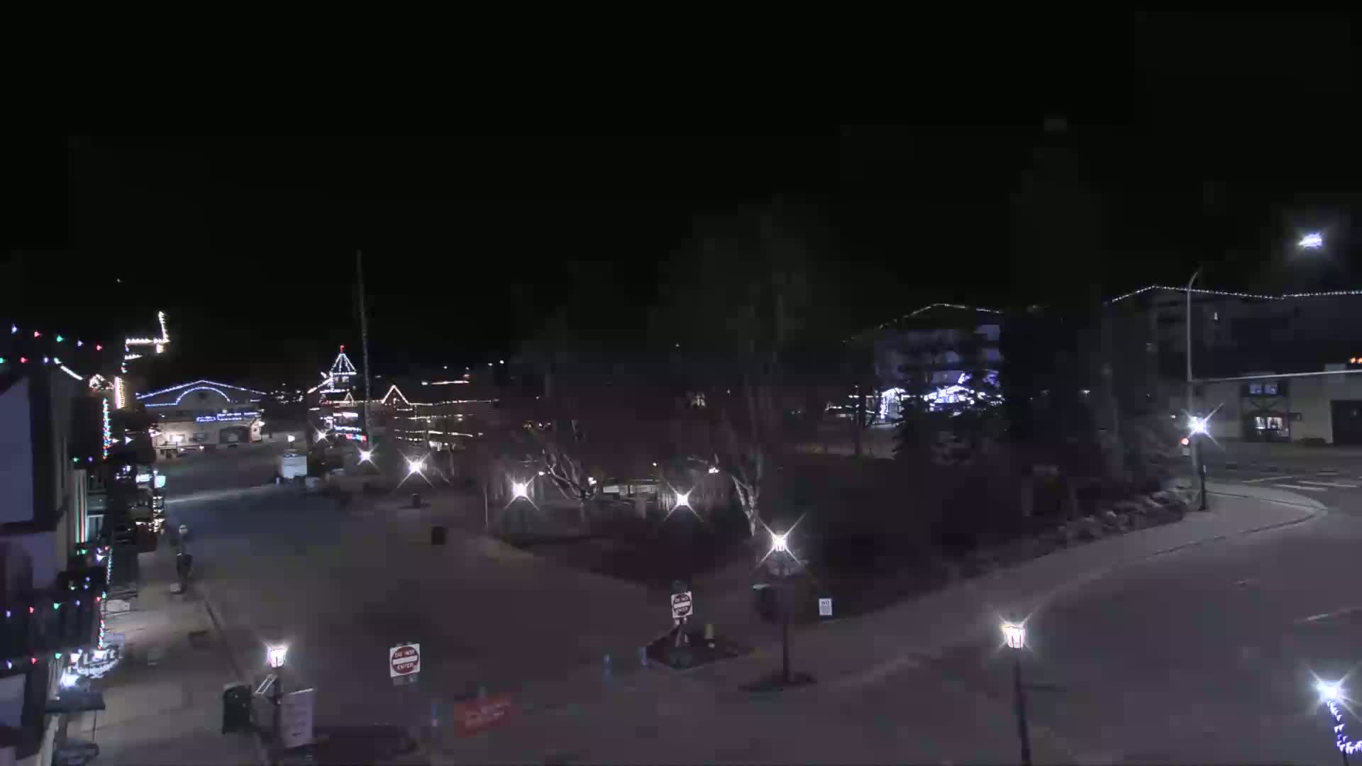 A nighttime view of a town square with buildings decorated with lights, streetlights illuminating the mostly empty streets, and dark trees in the center.