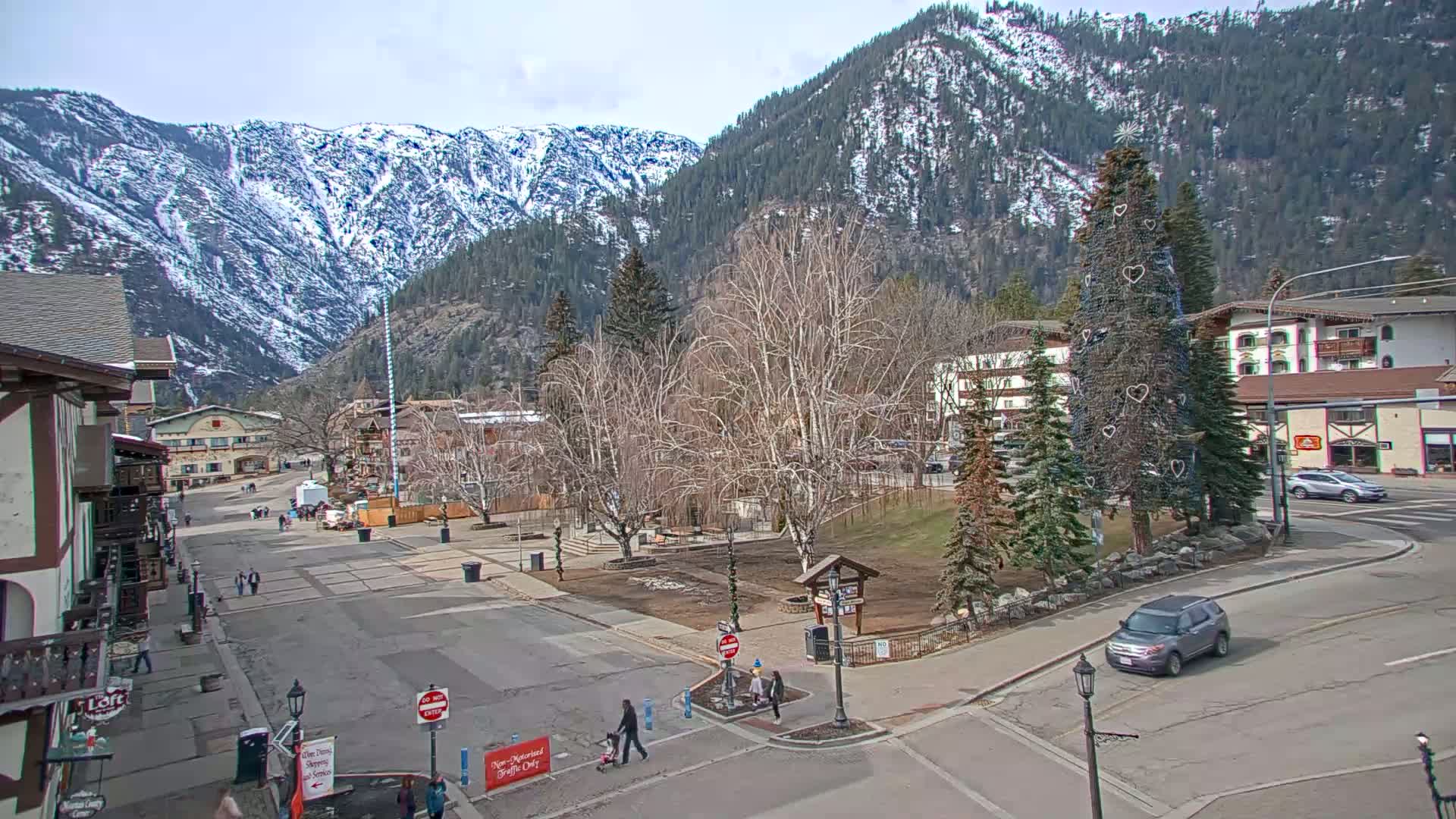 A town square with snow-capped mountains in the background under a sunny sky, featuring bare trees and a decorated evergreen.