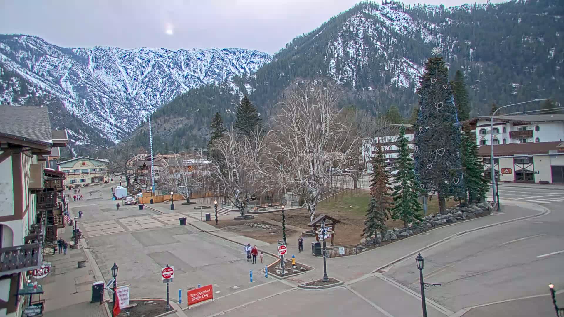 A mostly empty town square, with a decorated Christmas tree and snow-covered mountains in the background under an overcast sky.
