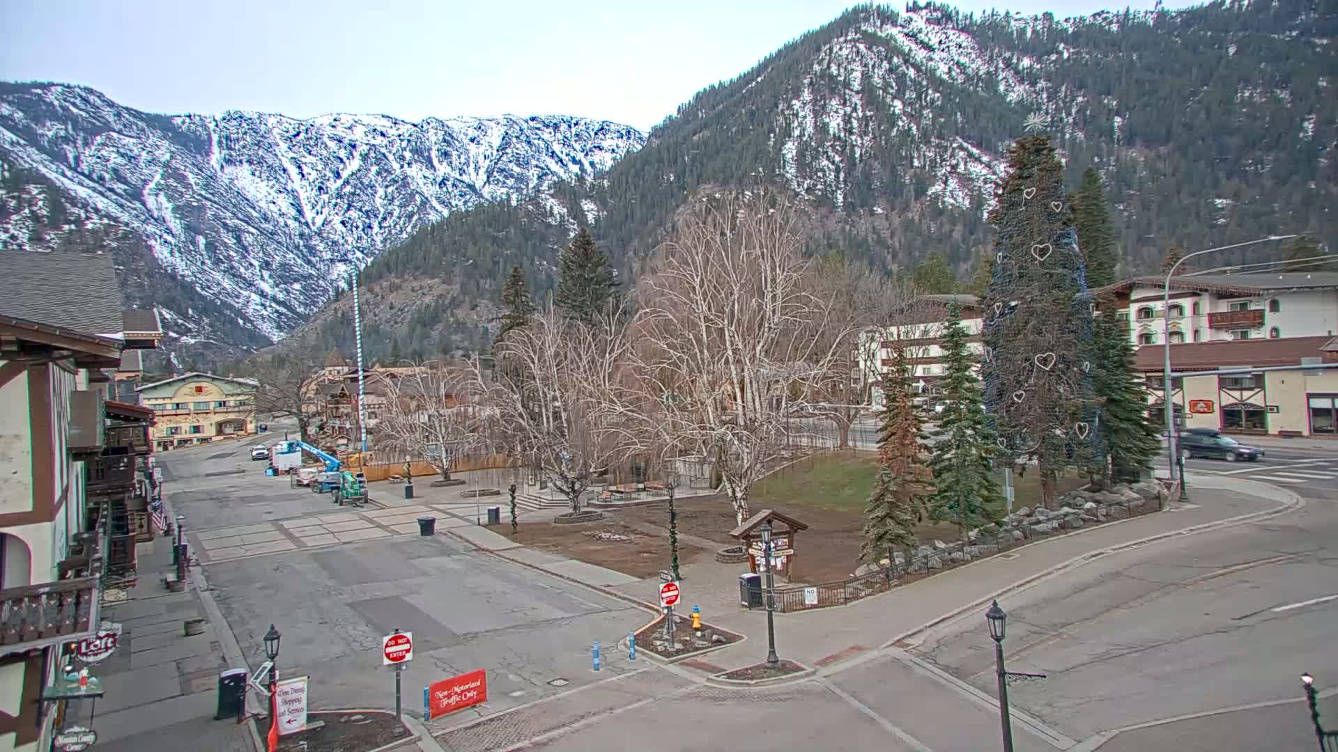 A mostly empty town square, with bare trees and a decorated evergreen tree in the center, is nestled in a valley surrounded by snow-capped mountains under a clear sky.