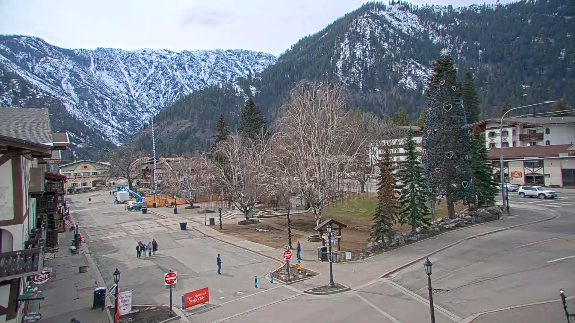 A mostly empty town square with snow-capped mountains in the background under a cloudy sky, featuring a decorated evergreen tree in the center.