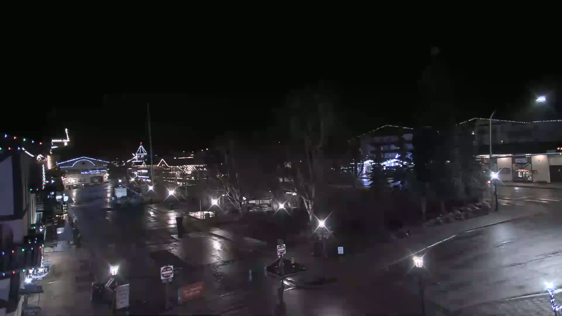 A wet, nighttime street scene shows a town square with several buildings decorated with lights.