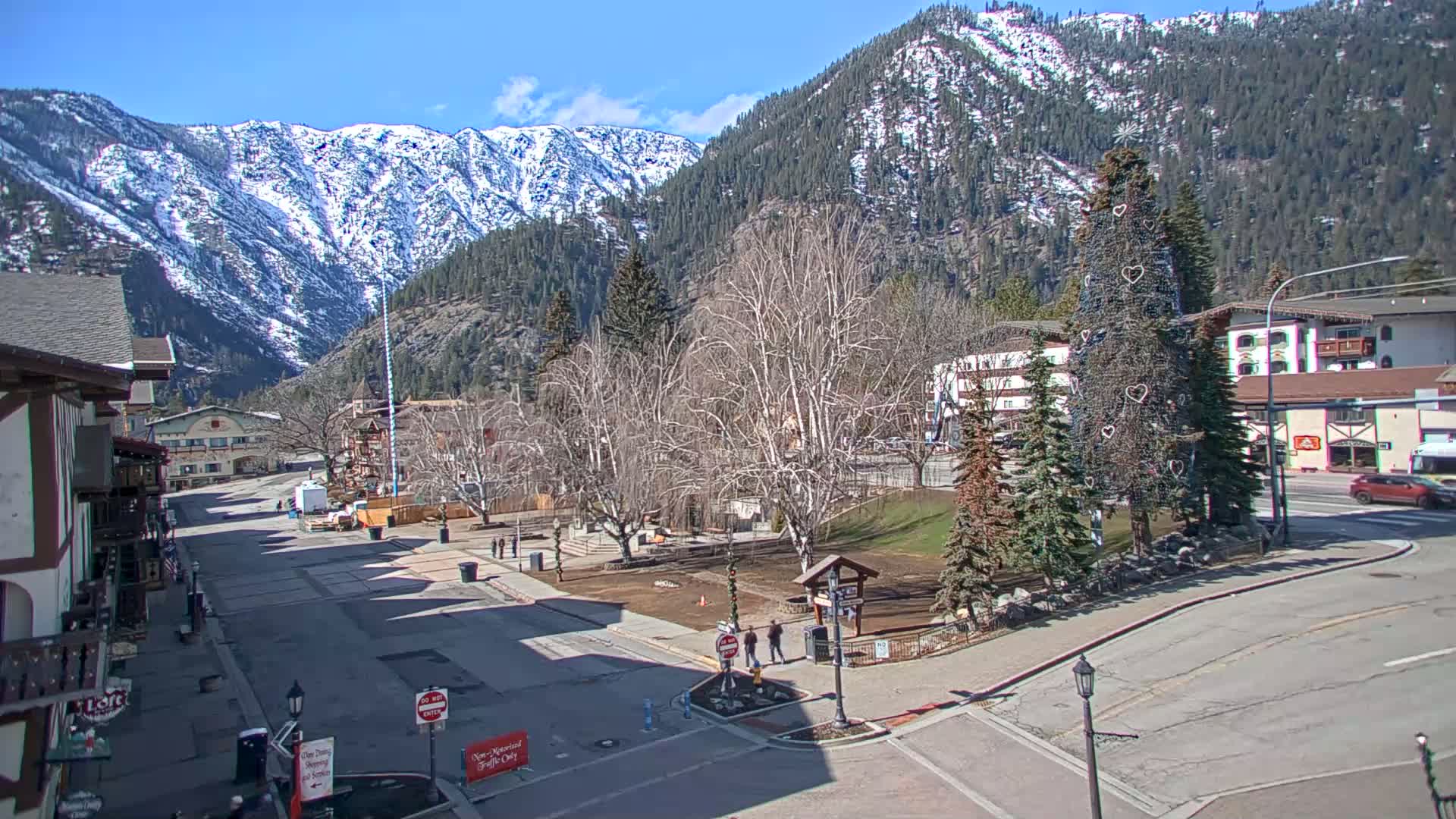 A mostly empty town square sits beneath snow-capped mountains on a sunny day, with a decorated evergreen tree in the foreground and bare deciduous trees surrounding a small park.