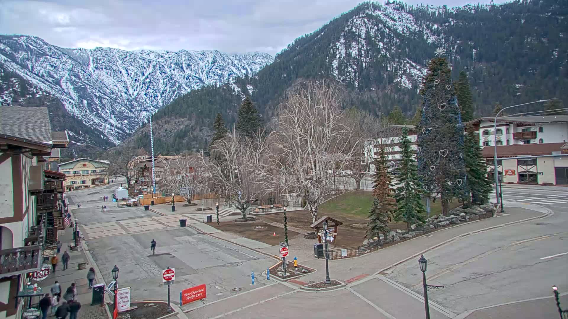 A mostly empty town square, surrounded by buildings and snow-capped mountains under an overcast sky.