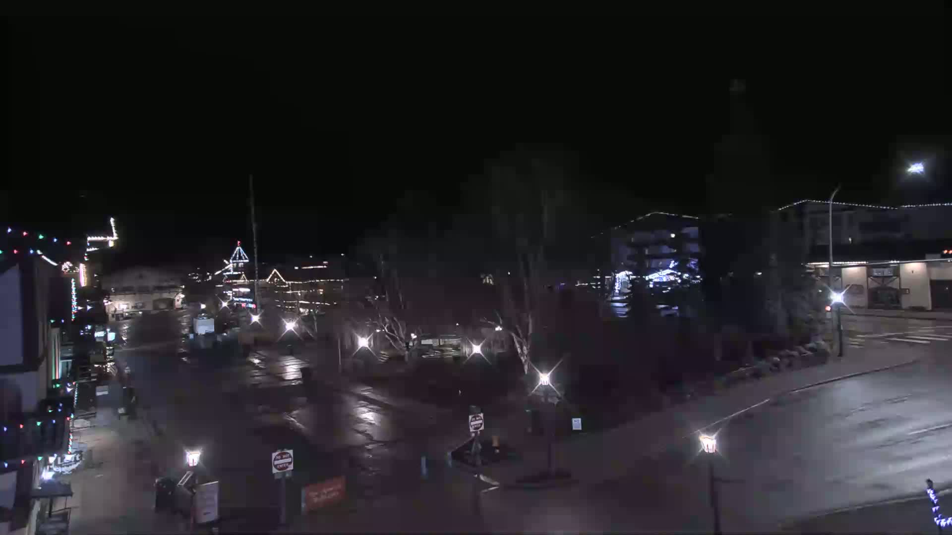 A nighttime view of a town square with wet streets, sparsely lit buildings decorated with Christmas lights, and a few cars.