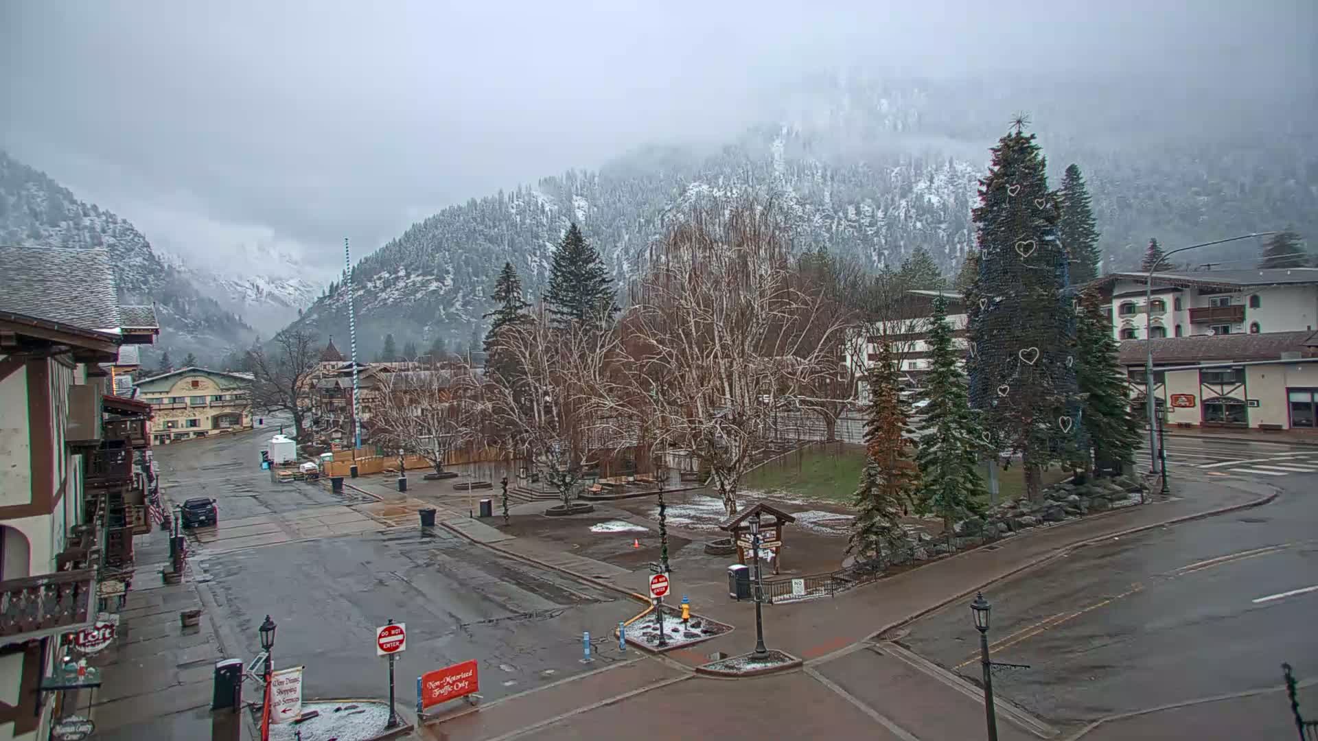 A snow-covered mountain town square with bare trees and a decorated evergreen tree is visible under an overcast sky.