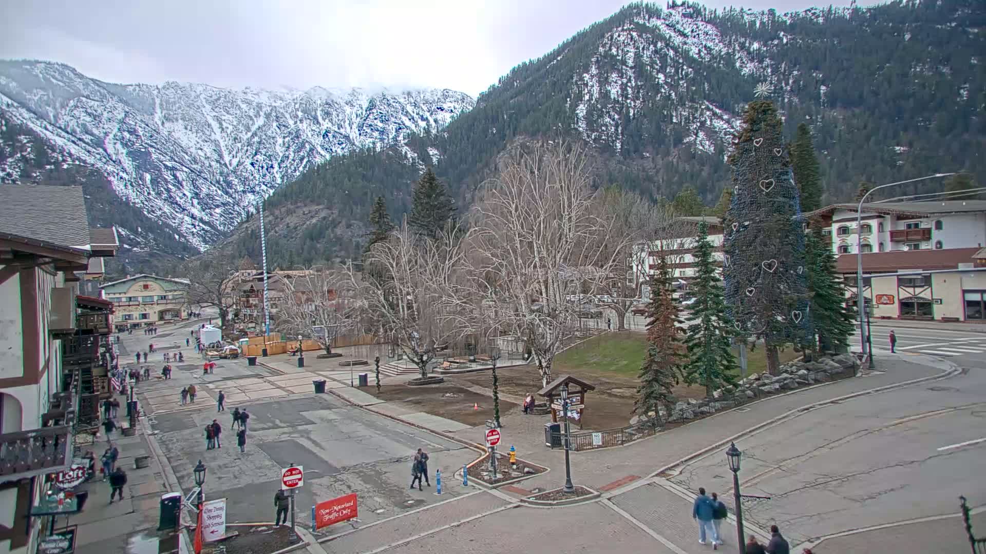 A snow-dusted mountain range overlooks a town square with people walking, bare trees, and a decorated evergreen tree in the overcast weather.