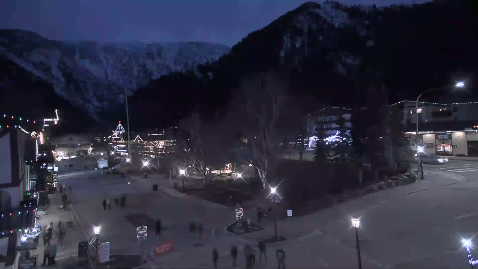 A nighttime view of a snow-dusted mountain town square, with pedestrians walking around illuminated buildings under a dark, clear sky.