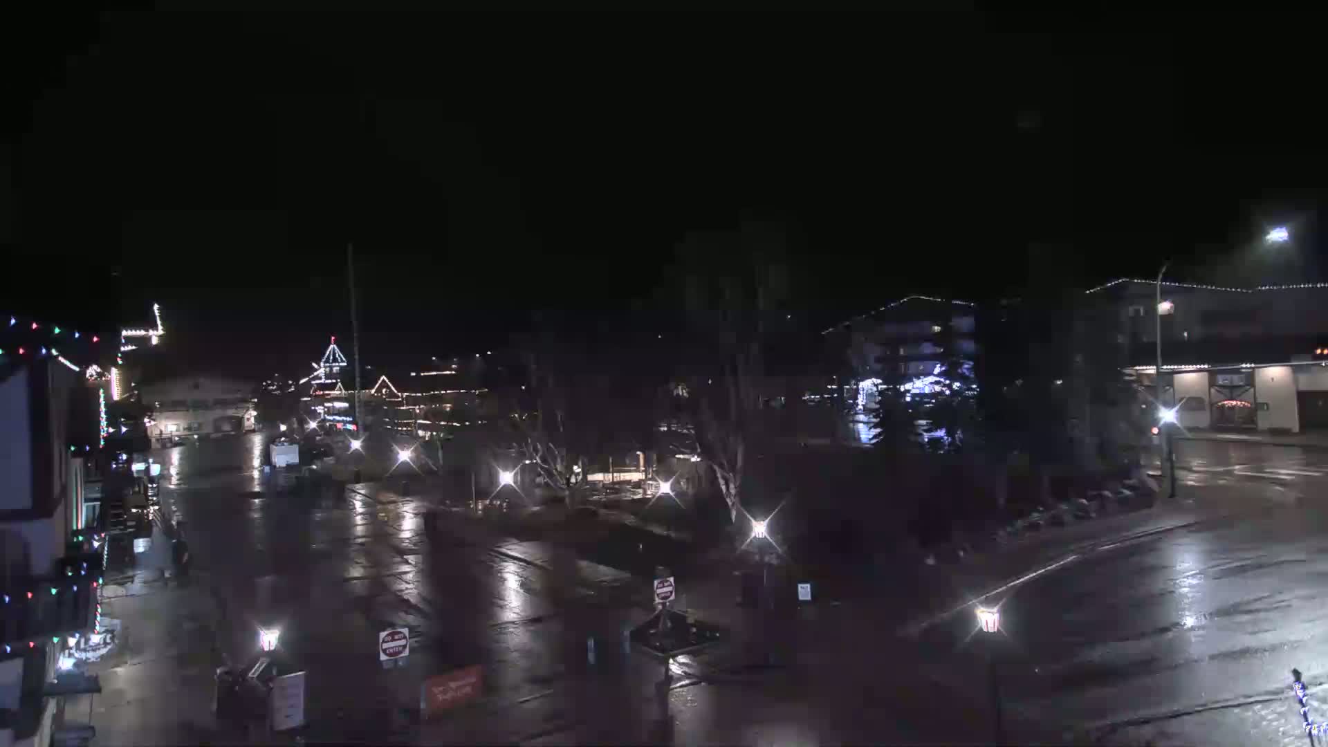 A nighttime view of a rain-slicked street in a town with buildings decorated with Christmas lights.