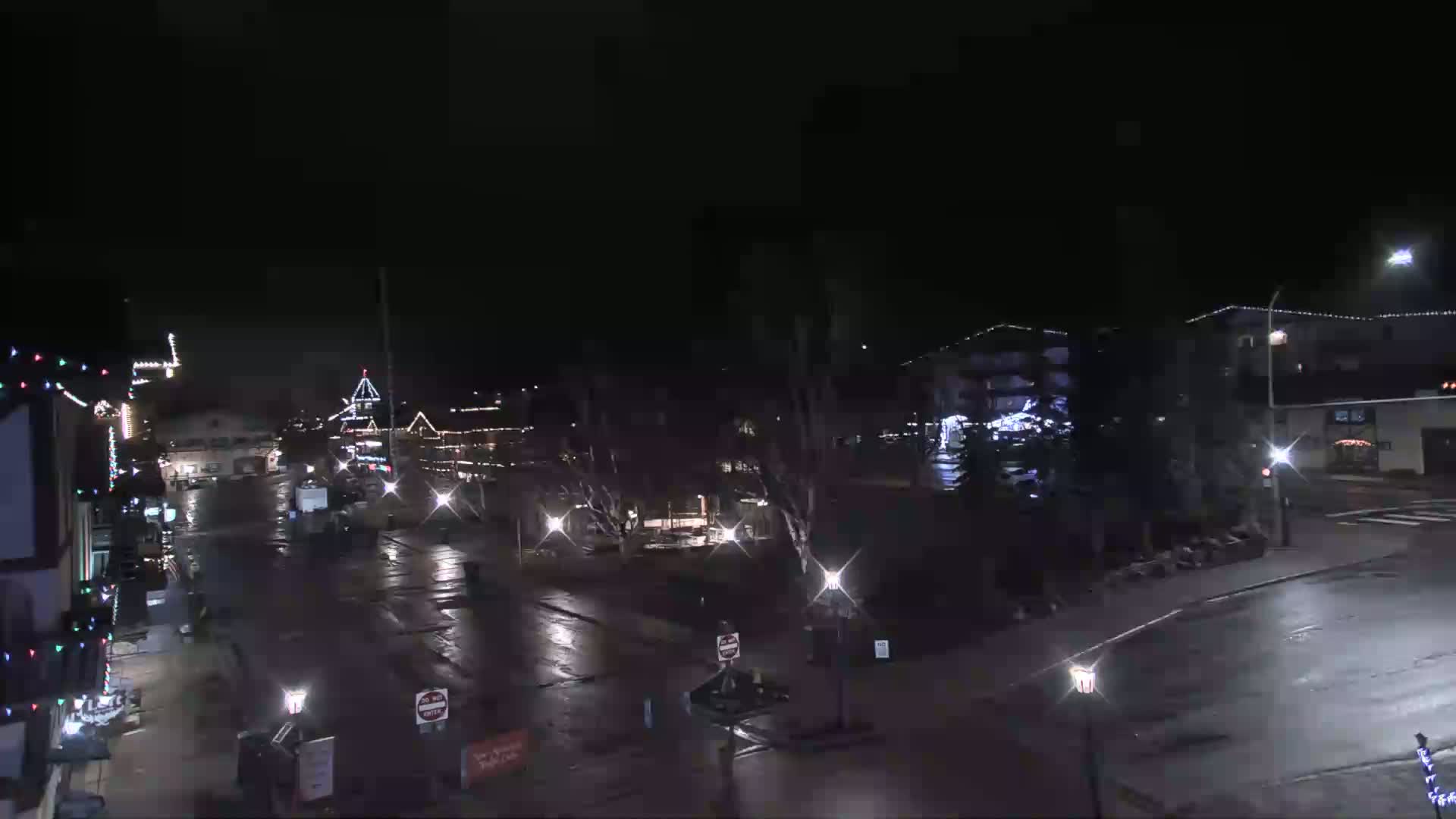 A nighttime view of a town square with wet streets, illuminated buildings decorated with holiday lights, and sparsely parked cars.