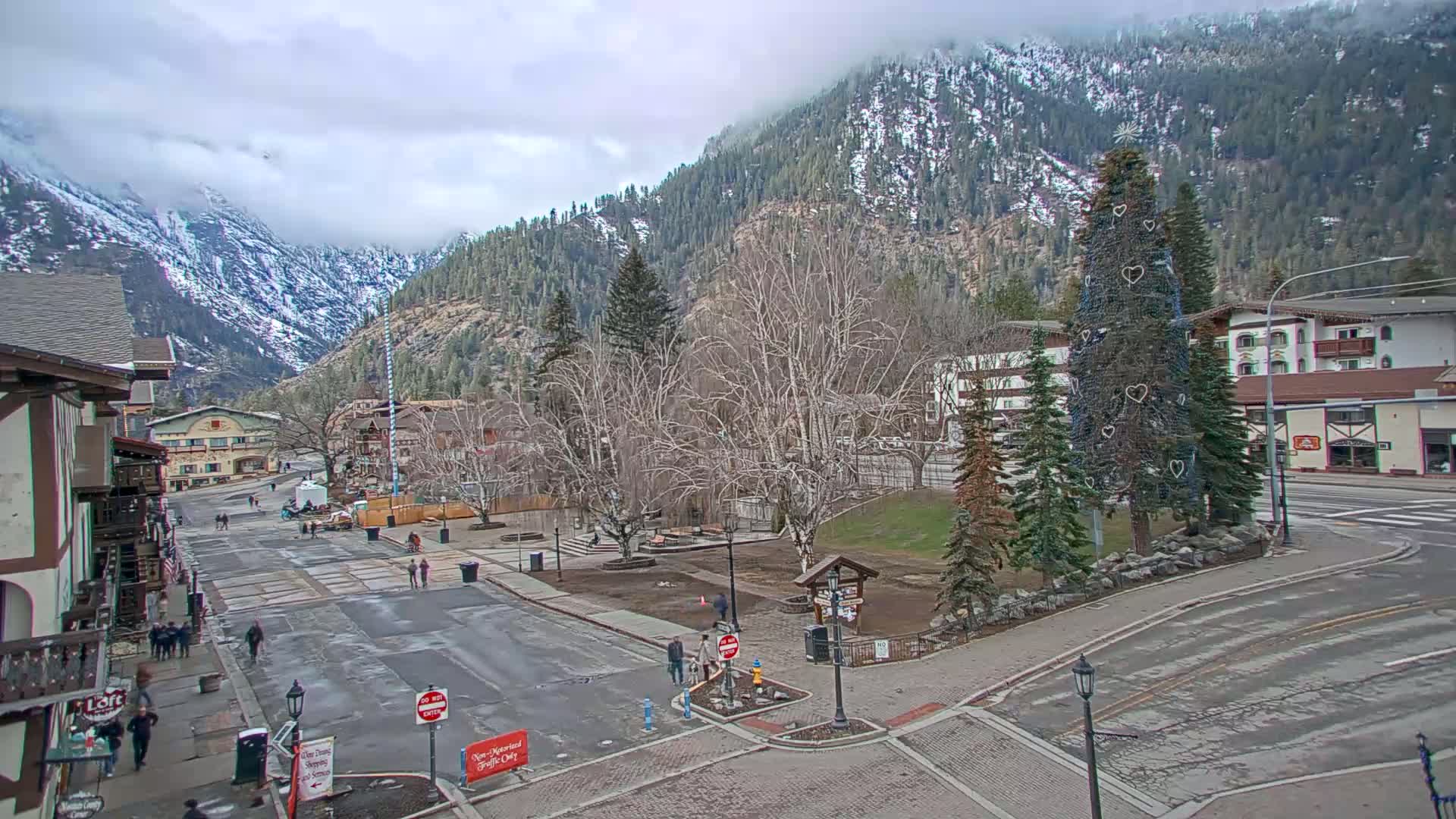 A town square with a decorated evergreen tree in the center, situated in a valley surrounded by snow-capped mountains under an overcast sky.