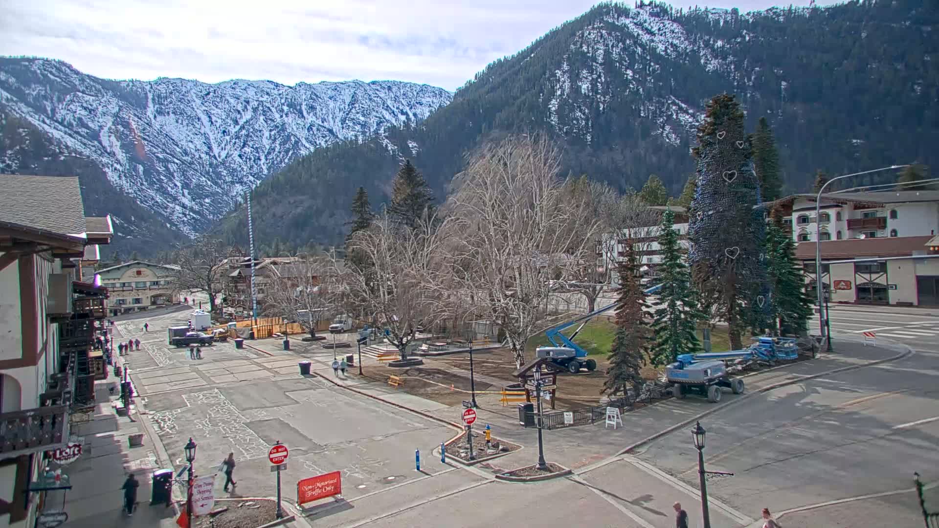 A town square with a large Christmas tree being worked on by a cherry picker, surrounded by buildings and snow-capped mountains under a partly sunny sky.