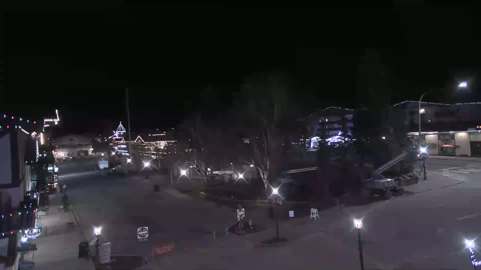 A nighttime view of a town square with Christmas lights on buildings and streetlamps illuminating a mostly empty street, under a clear, dark sky.