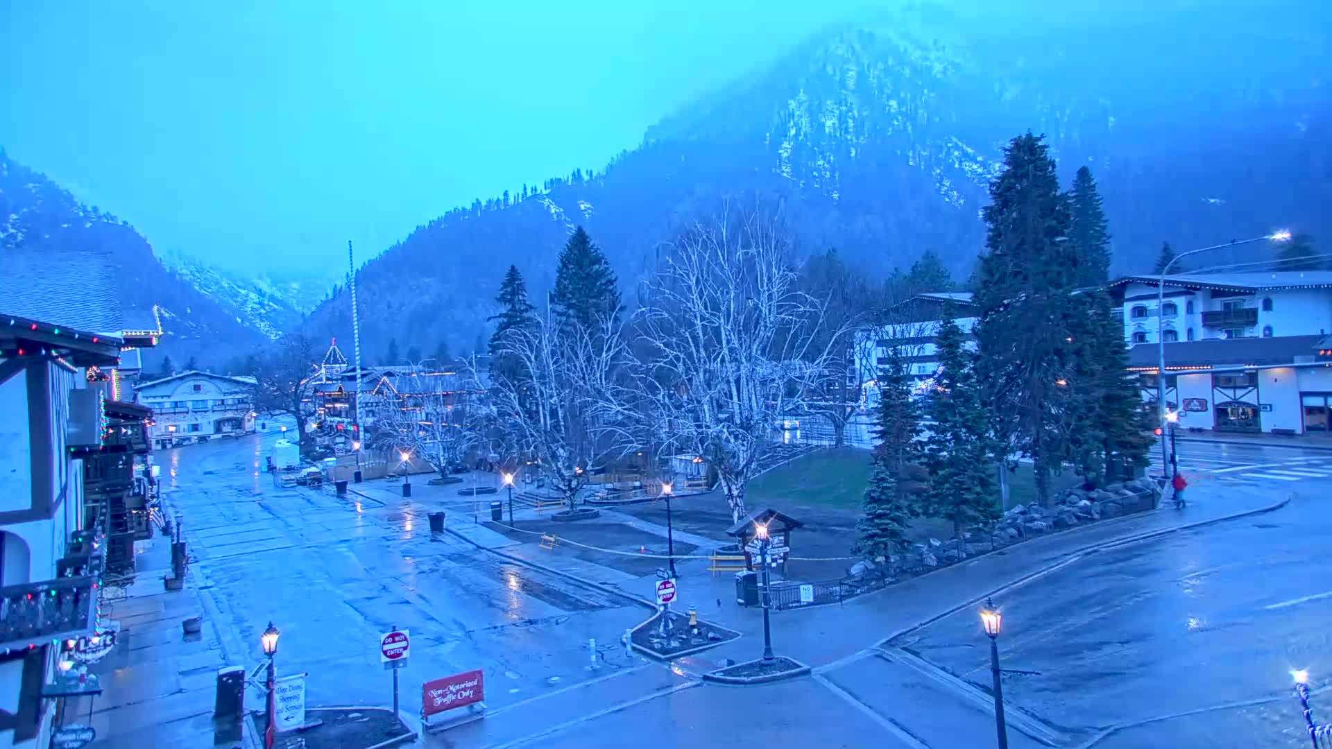 A wet, empty town square, nestled in a valley between snow-dusted mountains, is illuminated by streetlights under a blue, overcast sky.
