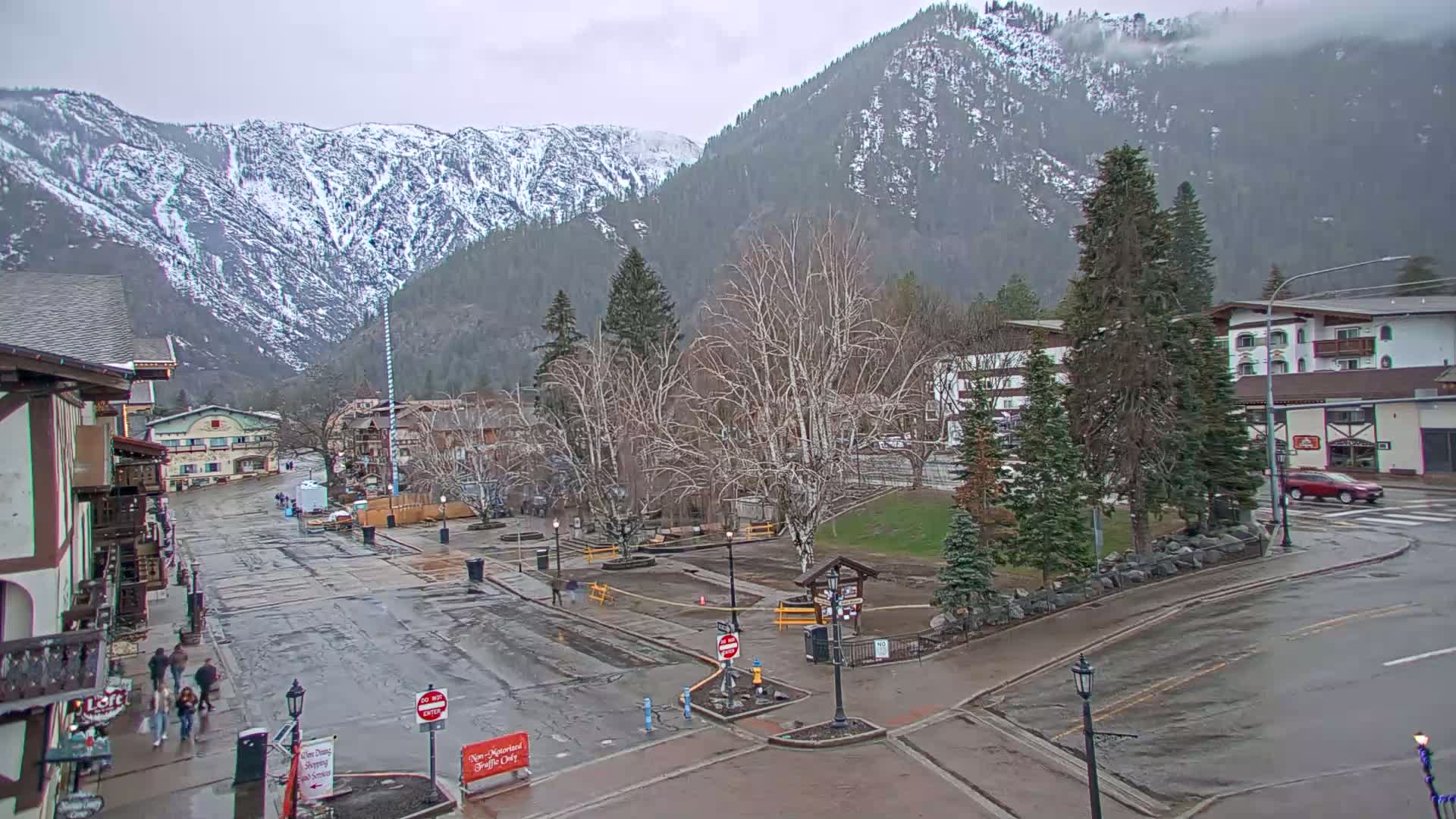 A wet town square with buildings and a park, situated in a valley surrounded by snow-capped mountains under an overcast sky.