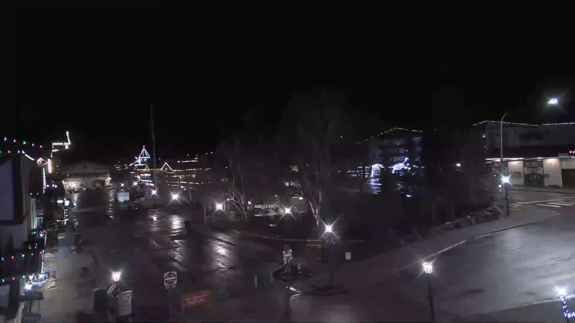 A nighttime view of a town square with wet streets and buildings decorated with holiday lights.