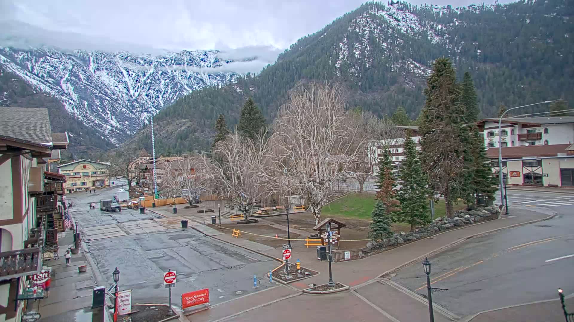 A mostly empty town square, under an overcast sky, is situated at the base of a snow-capped mountain range.