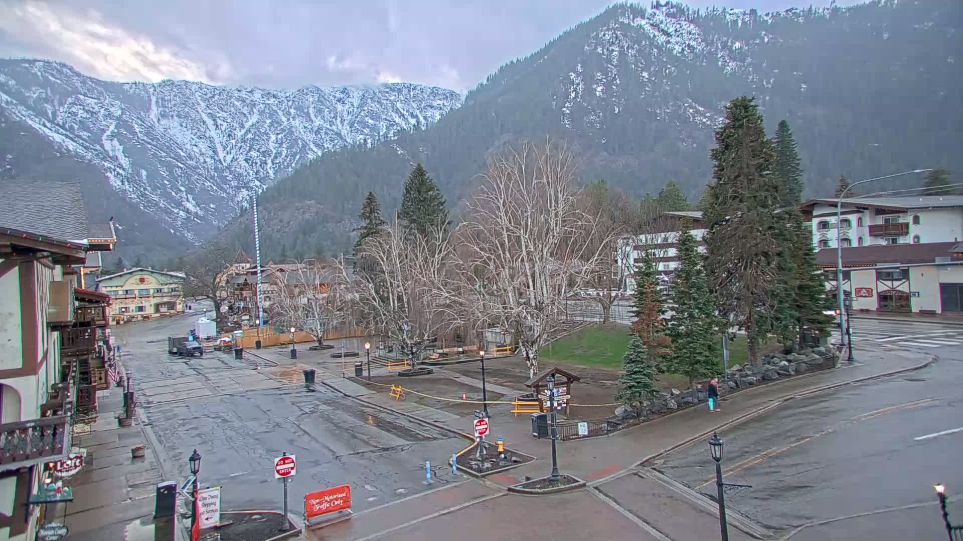 A wet, overcast town square sits at the base of snow-capped mountains, with bare trees and a few buildings visible.