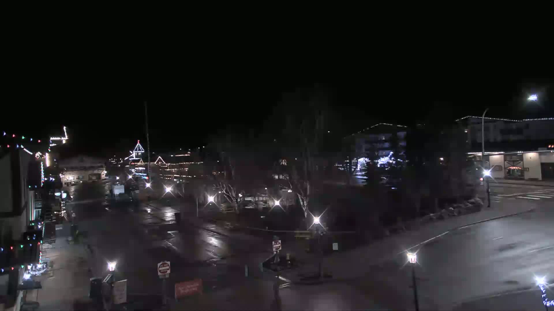 A mostly empty, wet street at night, lined with festively lit buildings and trees.