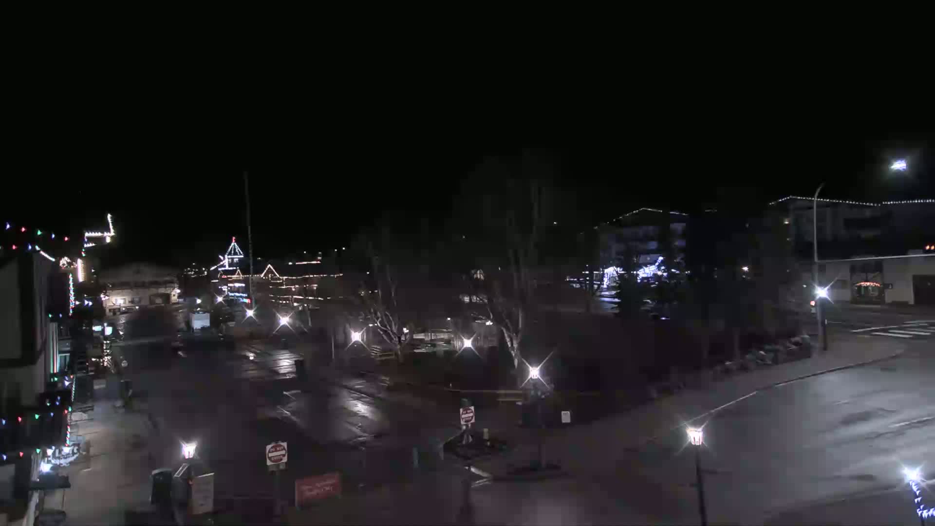 A mostly empty town square at night, with some buildings decorated with Christmas lights, is lightly wet from recent rain or snow.