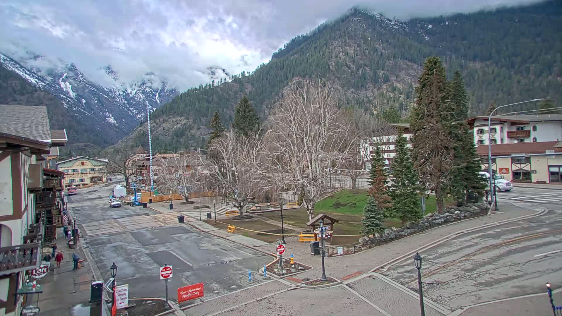 A partly cloudy day reveals a mountain town with a small park in the center of the street, surrounded by buildings and bare trees.