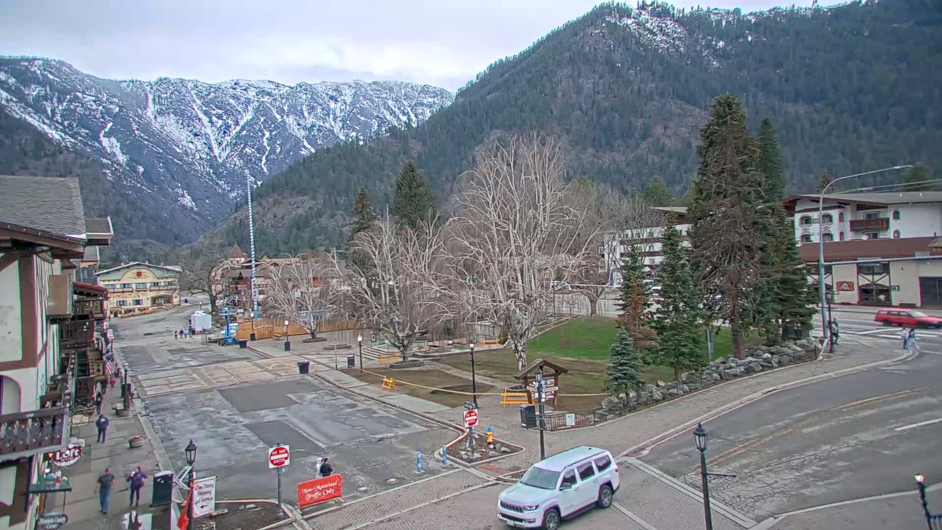 A mostly empty town square, surrounded by buildings and trees, sits in front of snow-capped mountains under a cloudy sky.