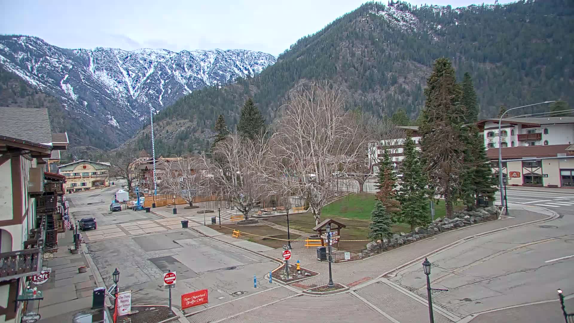 A mostly empty town square, with a few vehicles parked, sits at the base of snow-capped mountains under an overcast sky.