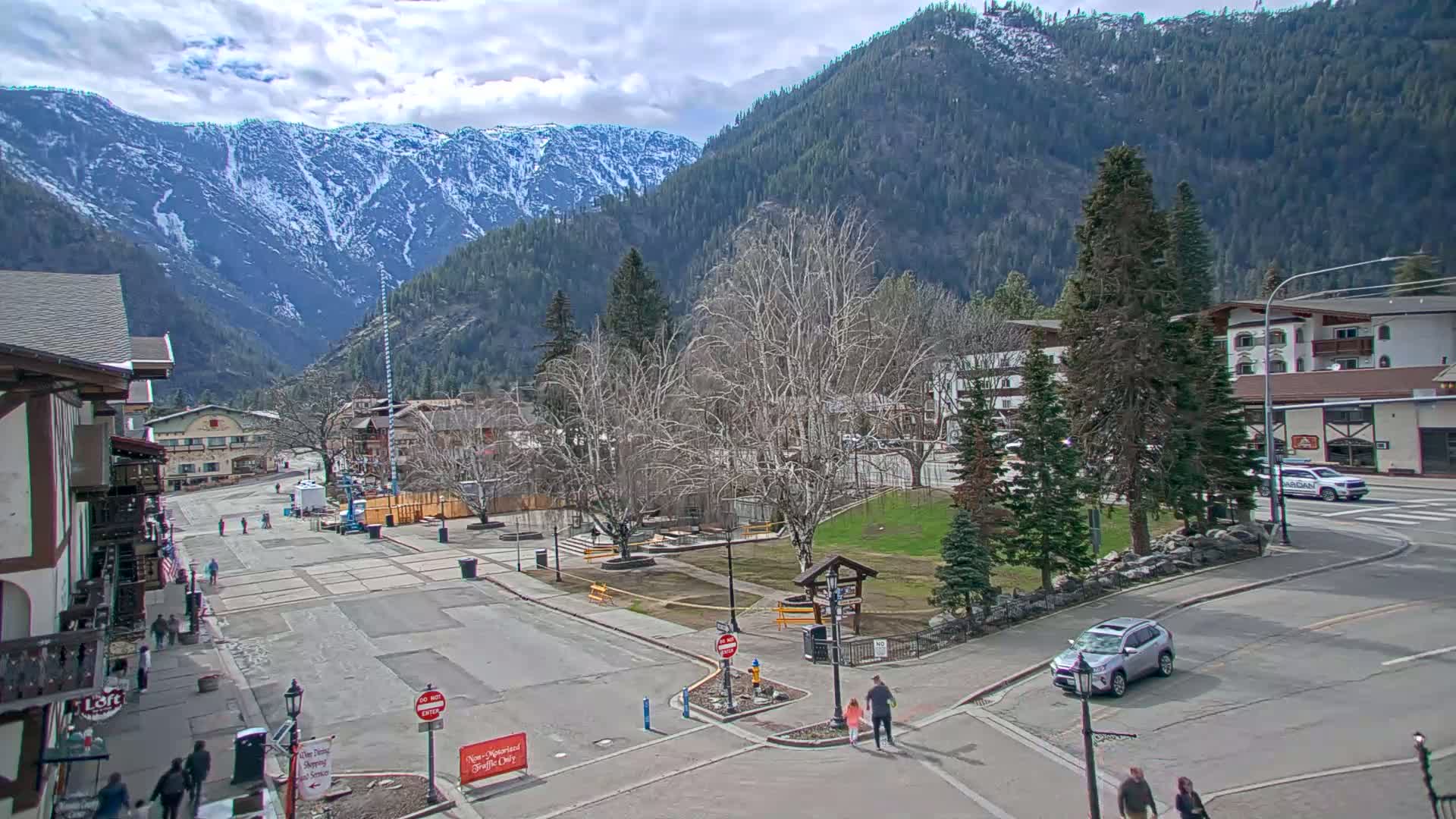 A mountain town square with snow-capped mountains in the background under partly cloudy skies, showing a few pedestrians and cars.