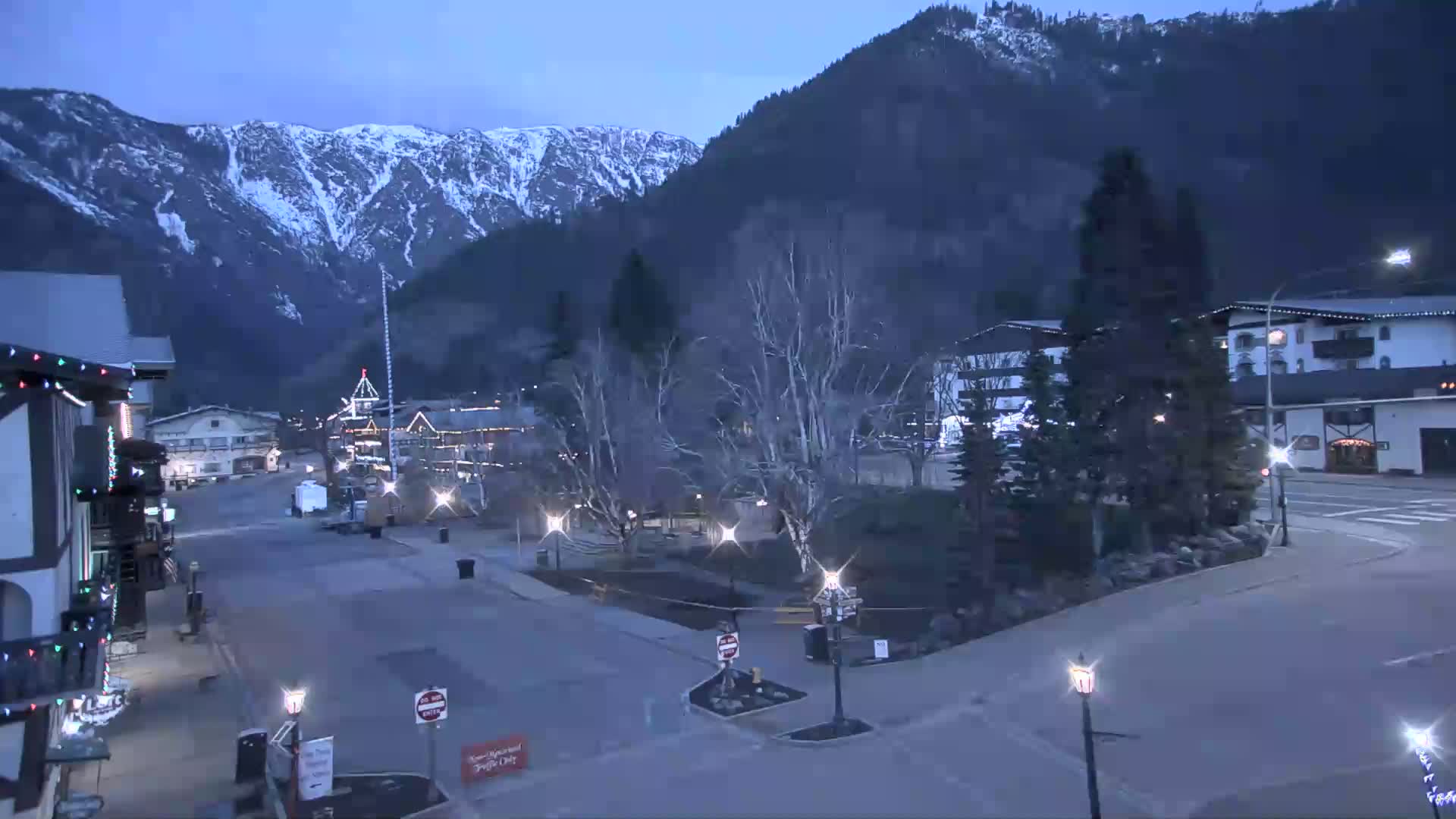 A mostly empty town square at twilight, nestled at the base of snow-capped mountains under a clear, dark sky.