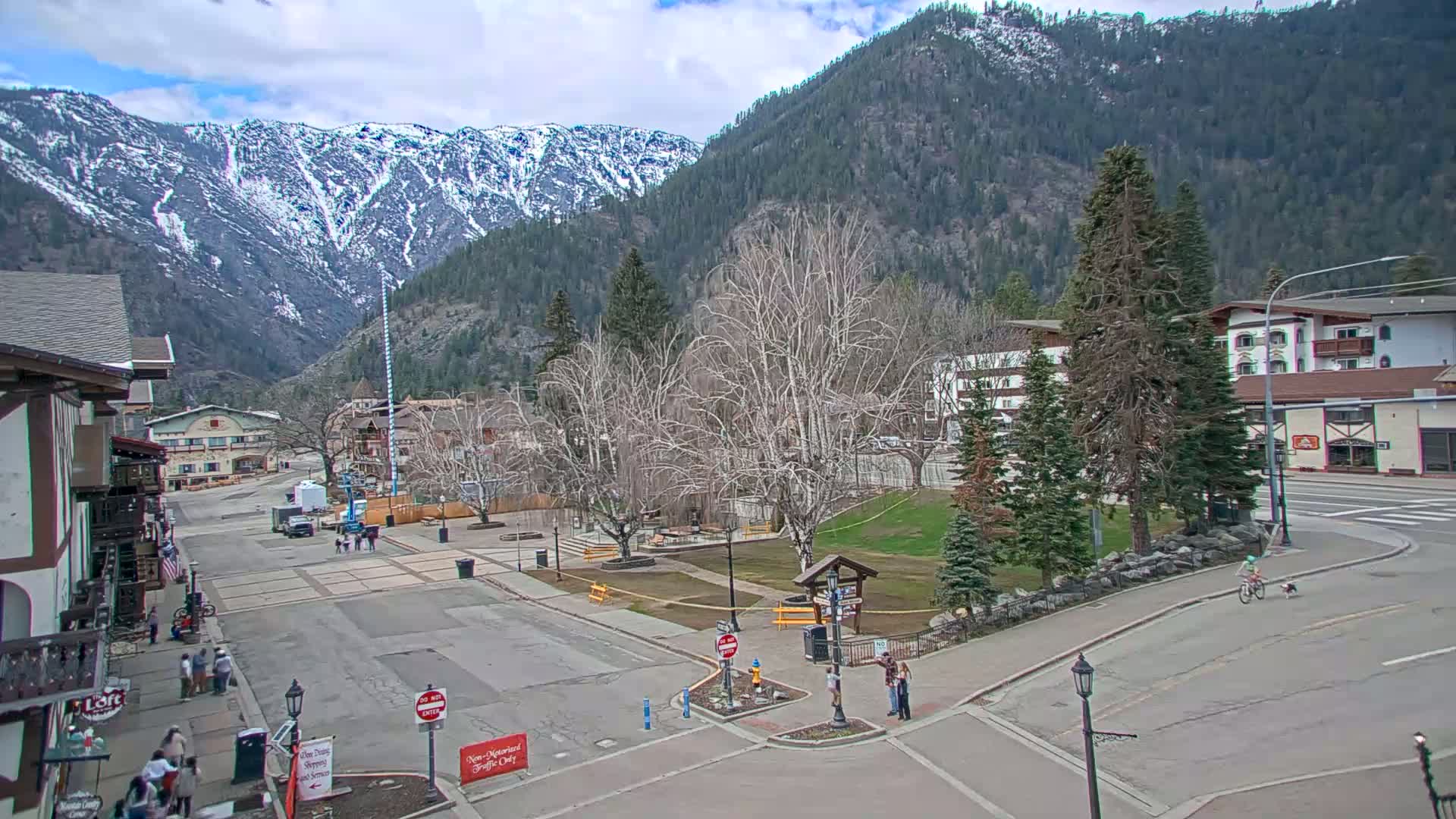A mostly sunny day in a mountain town square with snow-capped mountains in the background, bare trees, and a few pedestrians.