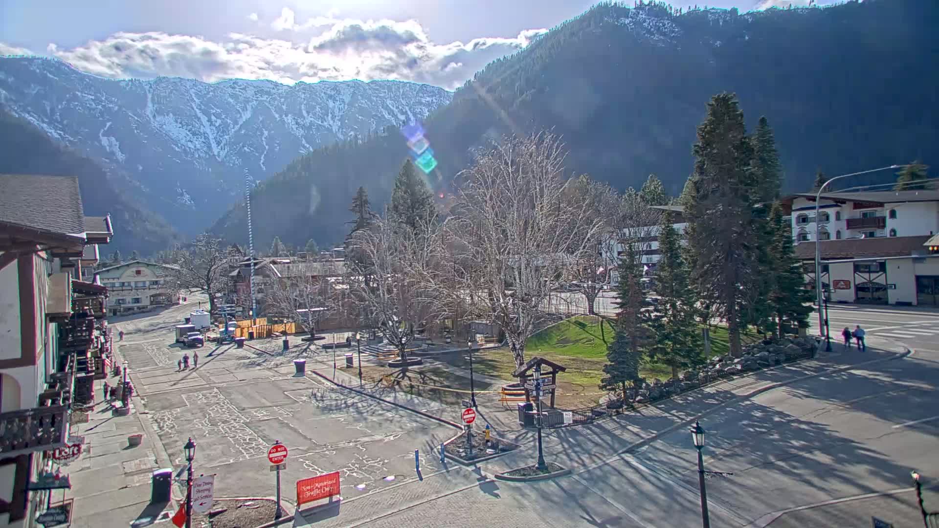 A sunny day reveals a snow-capped mountain range behind a town square with a few people walking and parked cars visible under mostly bare trees.