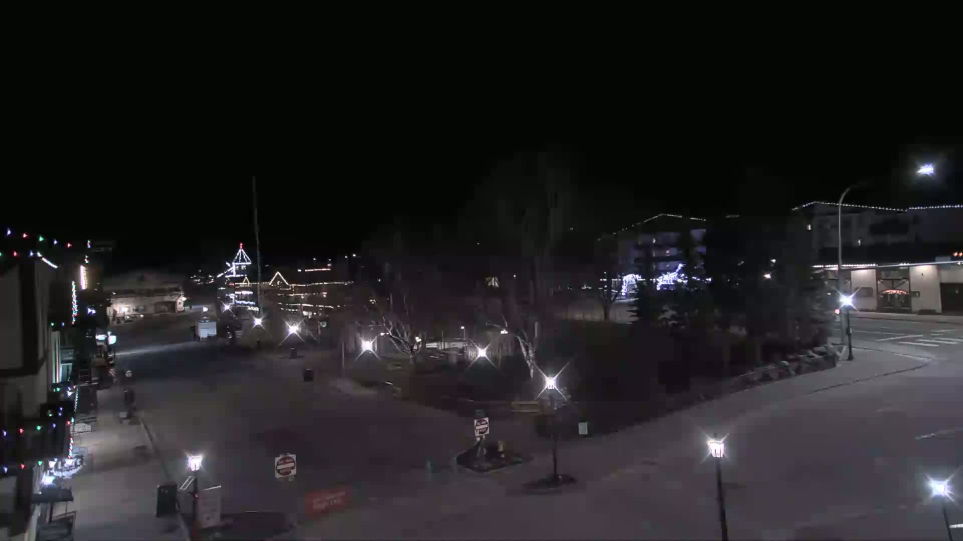 A mostly empty town square at night, illuminated by streetlights and building lights, with some sparsely-leafed trees.