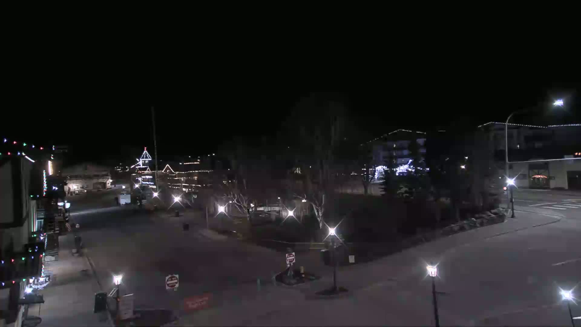 A nighttime view of a snow-covered town square with buildings illuminated by streetlights and holiday decorations.