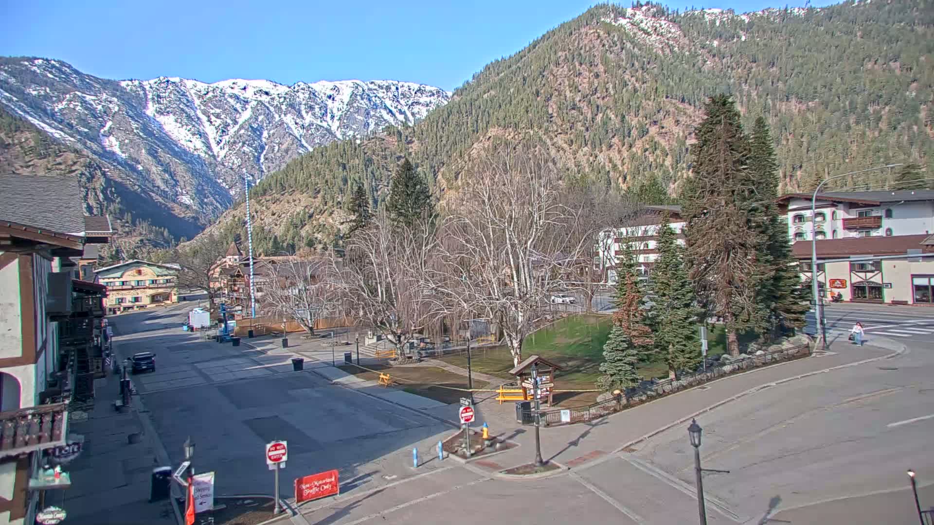 A mostly empty town square sits below snow-capped mountains on a sunny day, with bare trees and evergreen trees in the square and buildings lining the street.