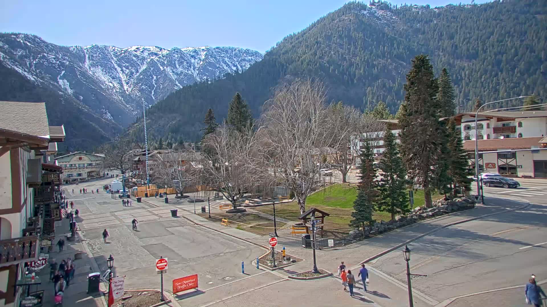 A town square with sparse pedestrian traffic is nestled between snow-capped mountains under a clear, sunny sky.