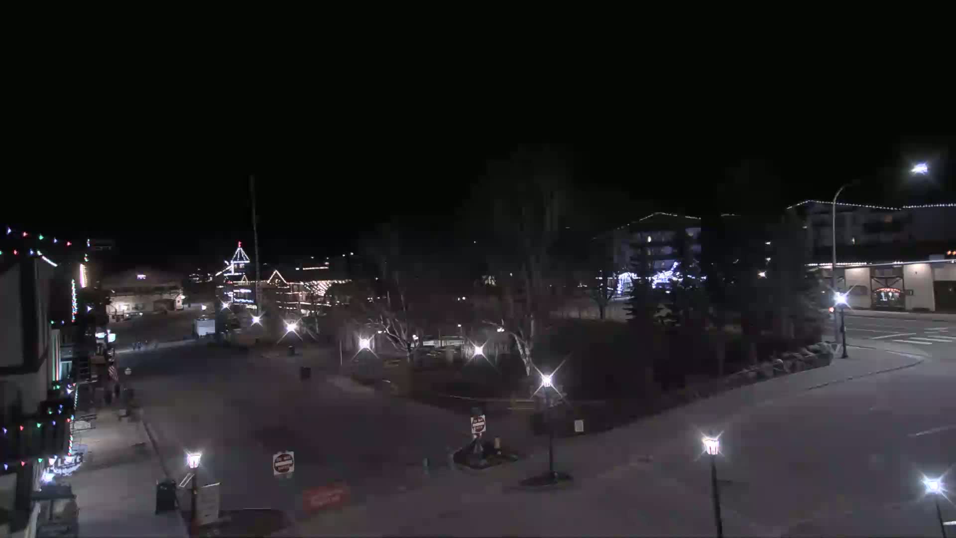A nighttime view of a town square with buildings, trees, and streetlights, under clear skies.