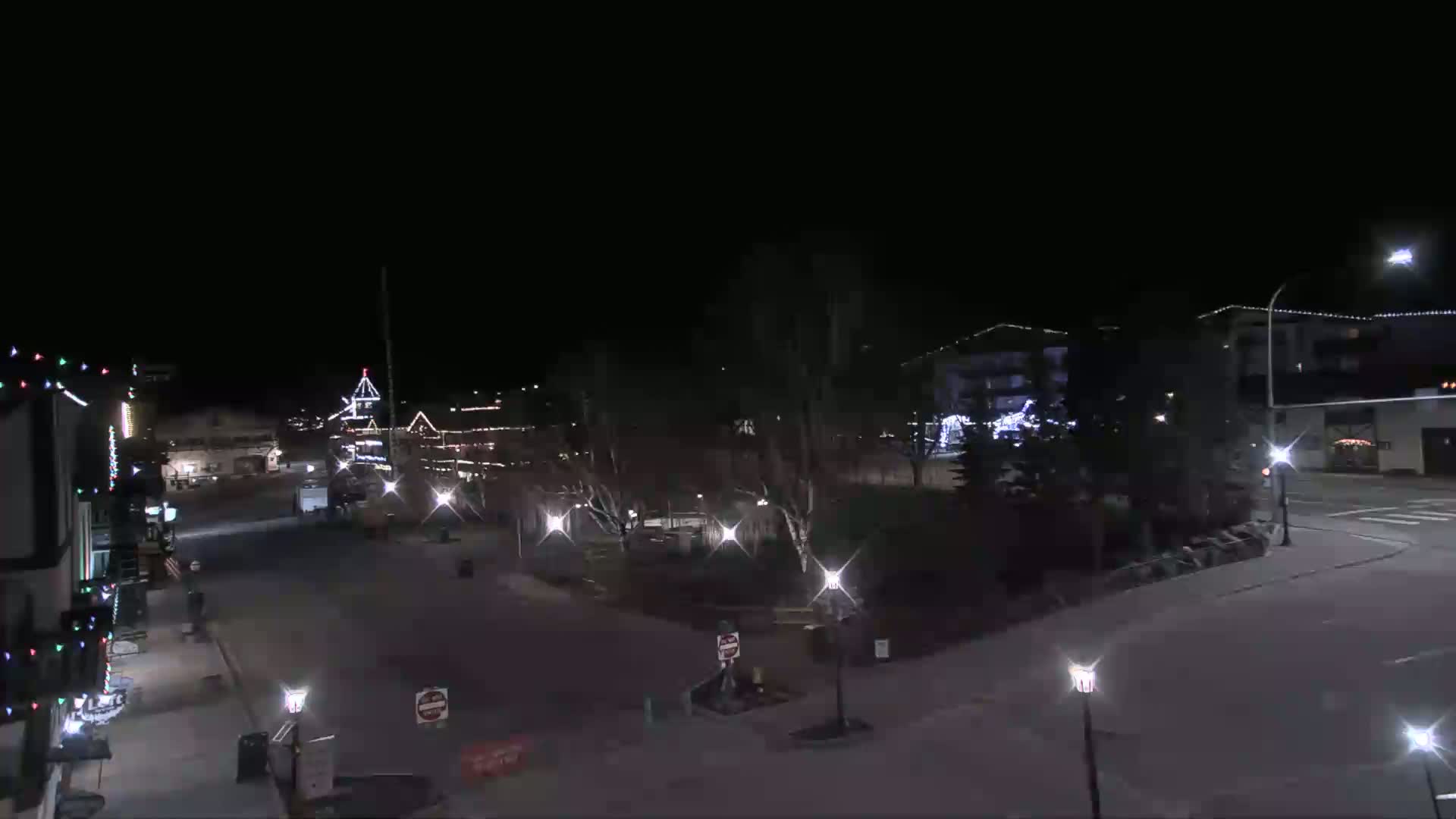 A nighttime view of a town square with buildings decorated with lights, trees, and streetlights illuminating the empty streets under a clear, dark sky.